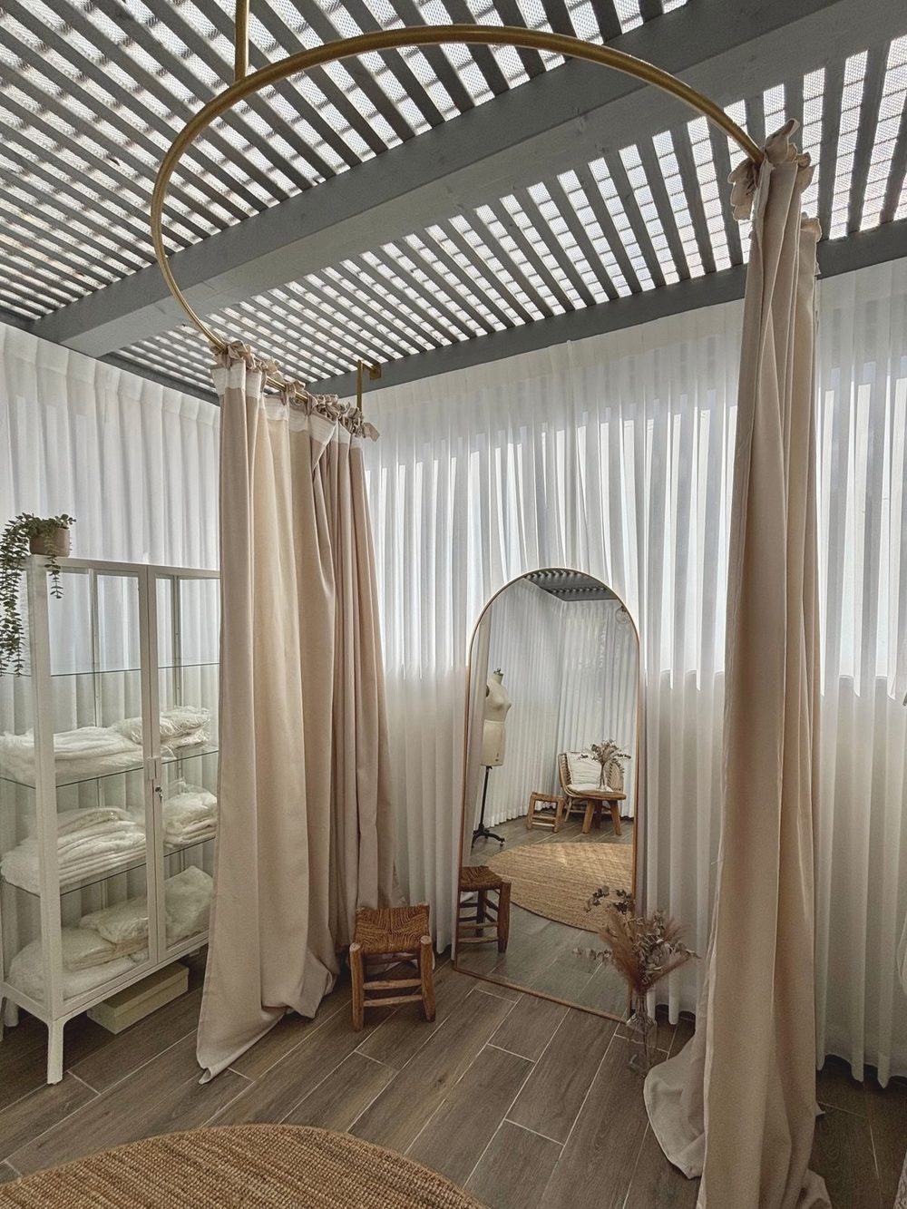 Fitting room in a bridal studio: a round mirror, sheer white curtains, a wooden stool and dresses hanging on a rail.
