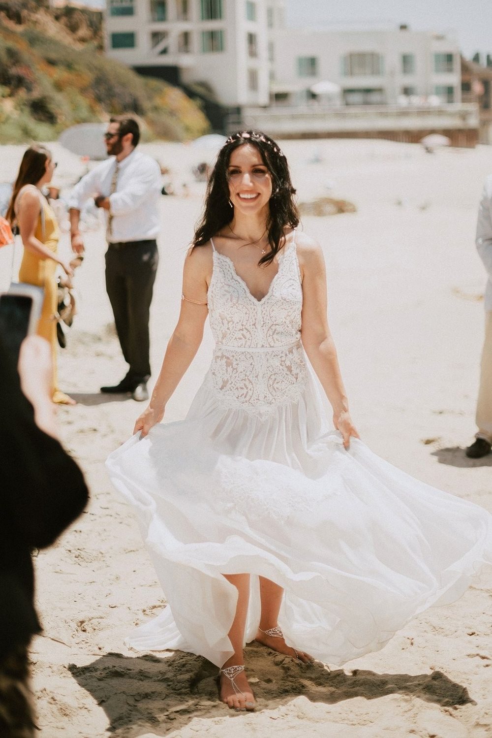 Smiling bride in an ivory lace and chiffon dress on a beach in full daylight, holding the skirt as it moves in the wind.