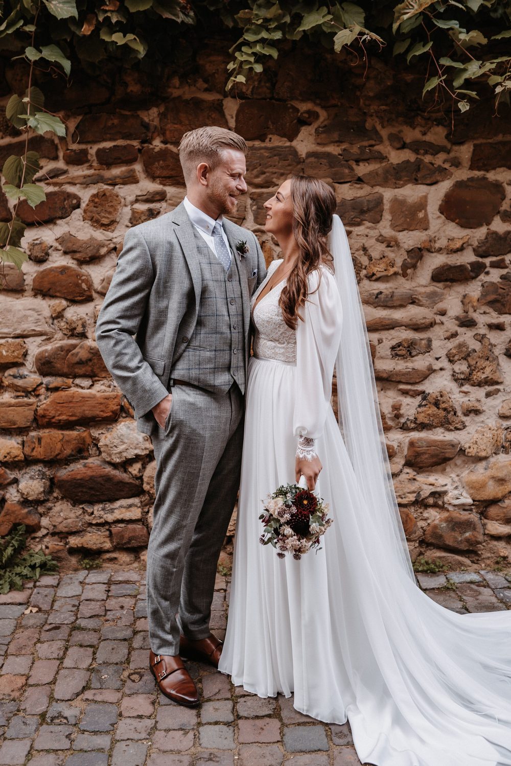 Bride in a plain wedding dress with a chapel-length train spread on cobblestones beside a stone wall, standing with her groom.