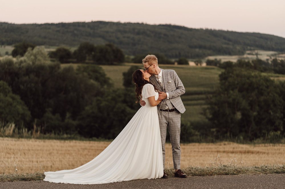 Bride in a plain A-line wedding dress with a long train and a groom in a grey suit, standing in open countryside at dusk.