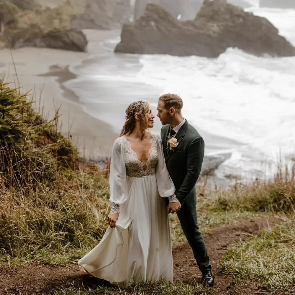 Bride in a long-sleeved chiffon and lace dress with her groom on a windy clifftop above a beach, the skirt blowing.