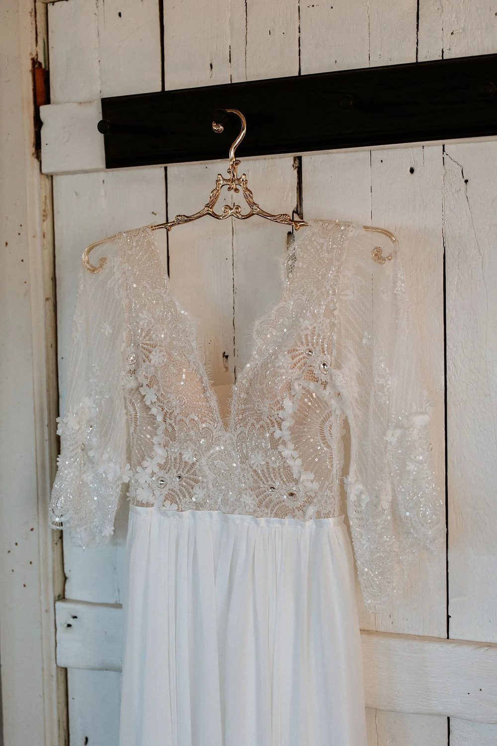 Long-sleeved lace wedding dress with a plunging neckline hanging on a wooden hanger against a white plank wall.