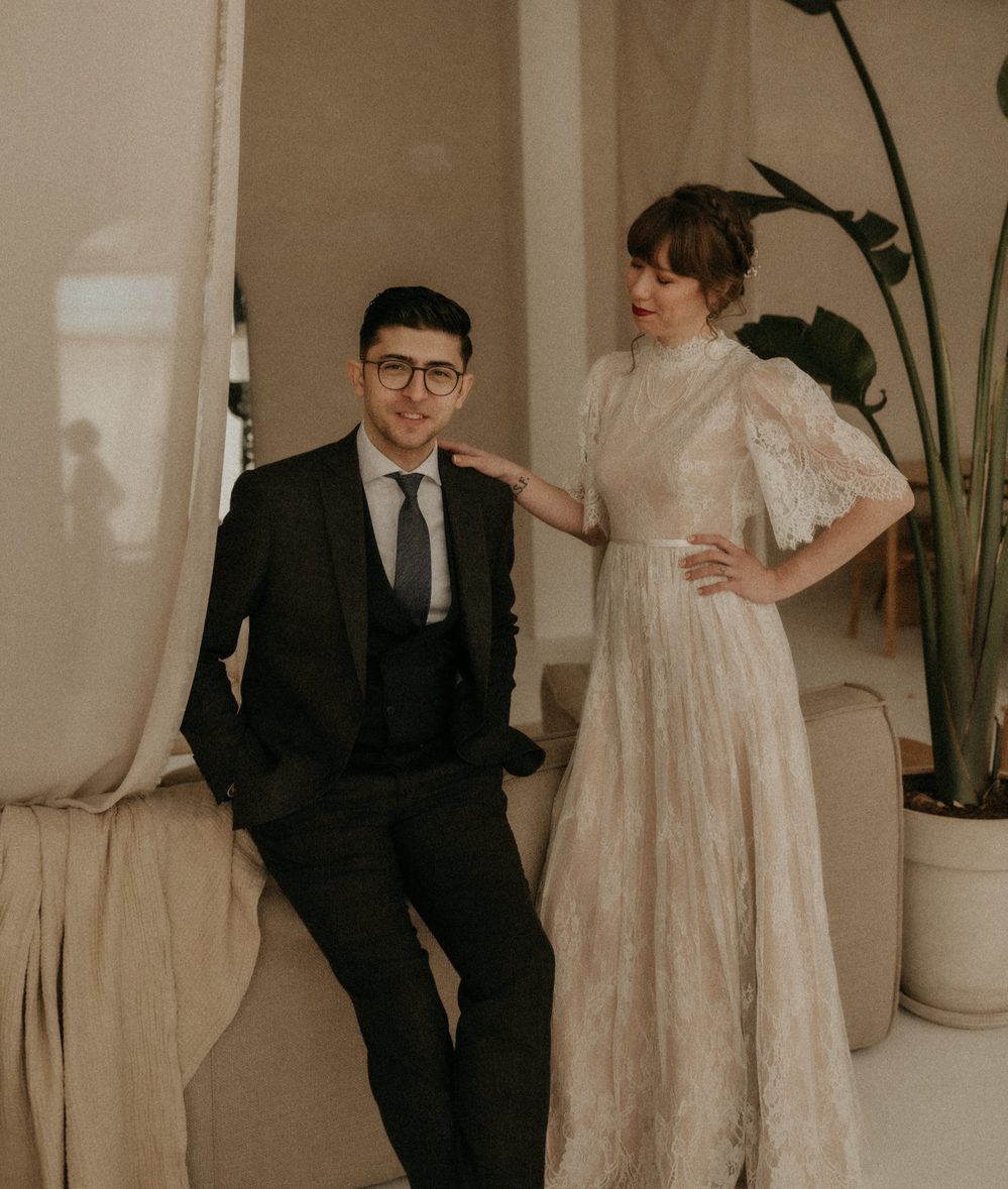 Bride in a vintage-style lace wedding dress with a high neck and short lace sleeves, standing beside her groom in a room with linen curtains and a palm.