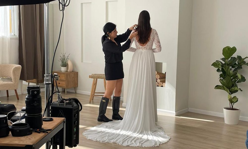 Bridal designer adjusting the shoulder of a long-sleeved lace wedding dress on a bride standing in a bright studio, with a camera on a tripod to one side.